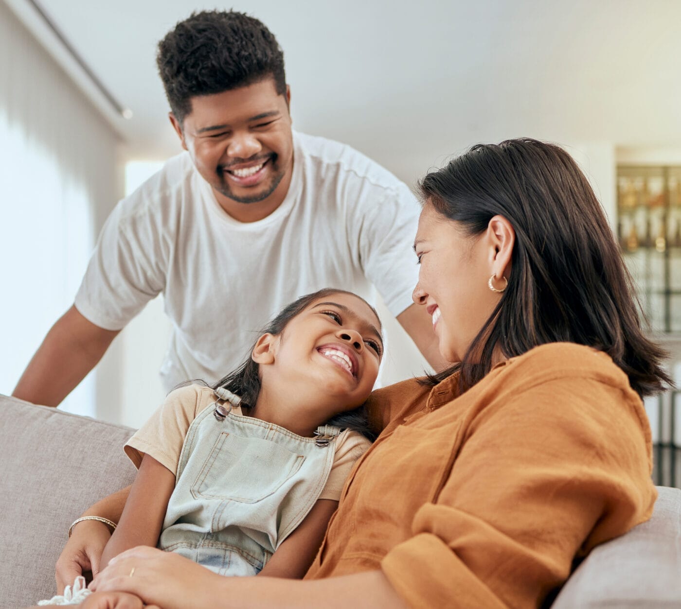 Family, mother and daughter hug with father standing at lounge sofa in Philippines home. Love, care and affection embrace of filipino mom bonding with happy child on couch in living room. Filipino family