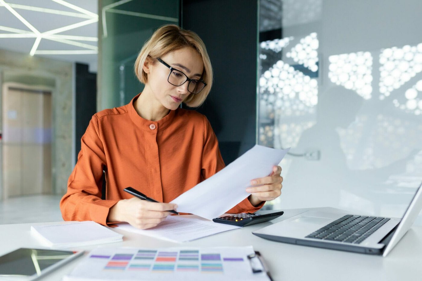 Serious and pensive business woman behind paper work inside office, female financier worker thinks about contracts and reports with charts and graphs, blonde successful woman uses laptop at work Employer filling out work visa paperwork