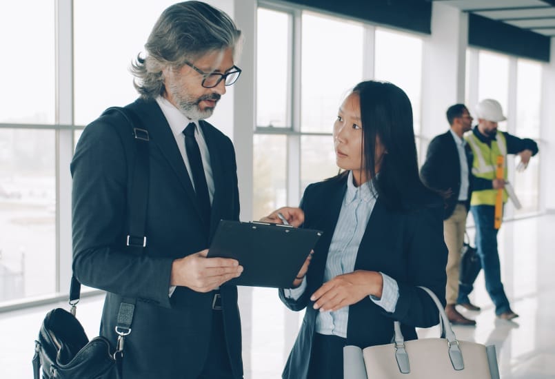 Multi-ethnic business partners man and woman are talking in modern building while workers in uniform are busy in background. 